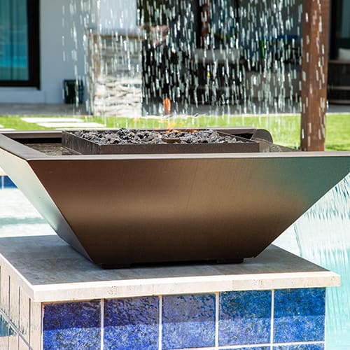 A modern, rectangular fire pit with a sleek, black metal design sits on a tiled base beneath a water feature. Water cascades down into a pool below, creating a serene and contemporary outdoor ambiance. The background shows part of a house and garden area.