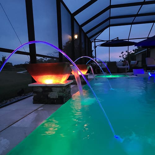 A luxurious backyard pool at dusk features illuminated fountains arching into the water. Warm, glowing fire bowls and ambient lighting create a serene atmosphere within a screened-in enclosure. Lounge chairs and outdoor seating can be seen in the background.