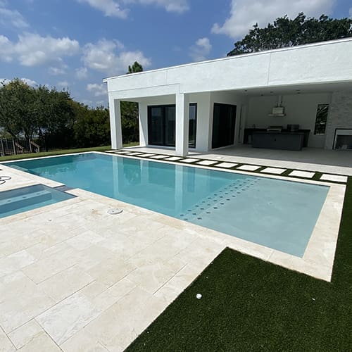 A modern backyard with a rectangular swimming pool next to a white one-story house featuring large glass doors. The pool area is surrounded by stone tiles and manicured grass, with a covered patio and outdoor kitchen visible in the background.