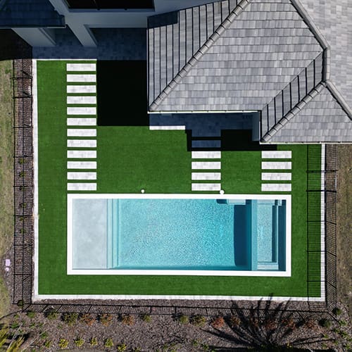 Aerial view of a modern rectangular swimming pool with clear water, surrounded by a manicured green lawn and paved stepping stones. The pool area is fenced and adjacent to a grey-roofed building. Shadows cast a symmetrical pattern on the lawn.