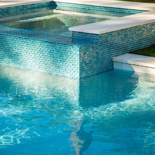 A sunlit swimming pool and spa with blue mosaic tiles. The pool's water is clear and shows a gentle ripple effect. The spa is elevated above the pool, with water cascading down the tiled wall into the pool below. The scene is peaceful and inviting.