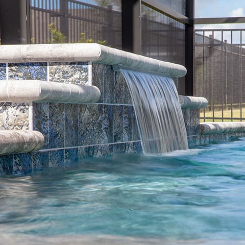 A close-up view of a swimming pool with a cascading waterfall feature. The water is clear and blue, flowing over a tiered stone structure into the pool below. The pool area is enclosed by a dark metal fence and a mesh screen, with a grassy area visible in the background.