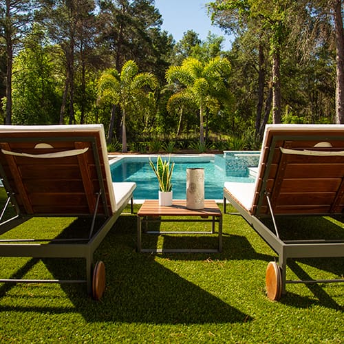 A serene poolside setting featuring two lounge chairs with white cushions and wooden accents on a vibrant green lawn. Between the chairs, there is a small table with a plant and cylindrical vase. The background showcases lush trees and palm trees.